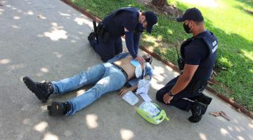 dois guardas estão agachados ao lado de modelo deitado no chão. Um dos guardar coloca o desfibrilador no peito do boneco. #paratodosverem