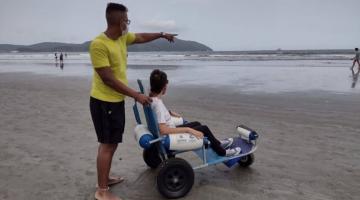 homem segura cadeira própria para entrar no mar onde está sentada uma jovem. ambos estão de costas para a foto.Ele aponta para o mar