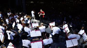 Músicos da Sinfônica de Santos estão em palco tocando diante de suas partituras em estantes. O maestro está ao fundo. Mais ao fundo, no escuro, se vê algumas luzes. #Pracegover