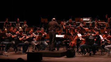 Orquestra sinfônica se apresenta em palco. Maestro está à frente do grupo, de costas para a imagem. #paratodosverem