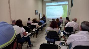 palestrante na frente da sala com telão ao fundo e pessoas assistindo #paratodosverem