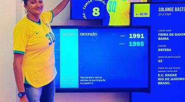 soré está em pé vestindo a camisa da seleção brasileira e apontando para um painel onde aparece a foto dela. #paratodosverem 