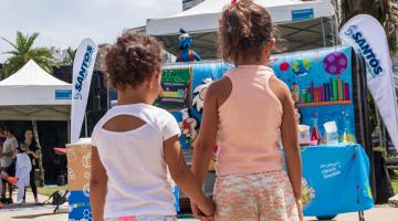 crianças estão de mãos dadas em frente a estande de brincadeiras ao ar livre. #paratodosverem 