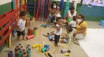 crianças uniformizadas brincam em espaço com  peças coloridas. Elas estão acompanhadas por mulheres que também estão sentadas no chão. #paratodosverem