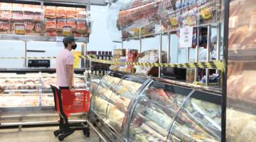 Homem está diante de balcão de carnes em supermercado. Ele está ao lado de um carrinho de mercadorias. O balcão está demarcado por faixa zebrada. #paratodosverem