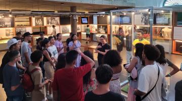 grupo de pessoas presta atenção em intérprete de libras que está diante deuma estátua do Rei do Futebol, no Museu Pelé. #paraatodosverem 