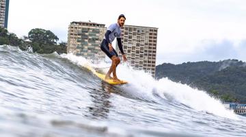 Atleta surfando na crista da onda em prancha longa com prédios e morro ao fundo. #paratodosverem