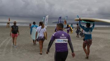 Alunos se dirigem ao mar carregando pranchas no alto da cabeça ou nos braços. O dia está nublado. #paratodosverem