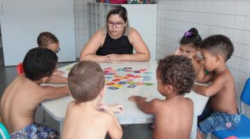 Crianças estão sentadas em torno de uma mesa. Elas estão jogando um jogo acompanhadas por uma mulher. #Pracegover