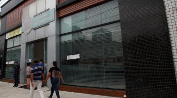 pessoas caminham ao lado de um prédio com parte da fachada em vidro #paratodosverem
