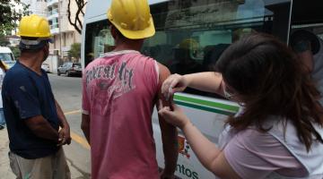 Mulher usando avental branco vacina homem que usa capacete de construção civil. Eles estão à frente de um veículo que tem o brasão da Cidade. #Pracegover