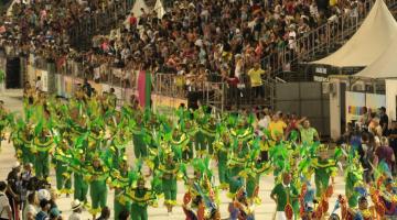 Desfile de escola de samba em passarela, com o público na arquibancada ao fundo. #Paratodosverem