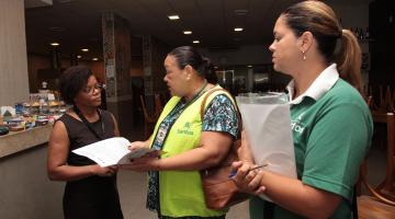 Agentes da Ouvidoria vestidas com colete e camiseta de identificação conversam com uma mulher em um estabelecimento comercial. #Pracegover