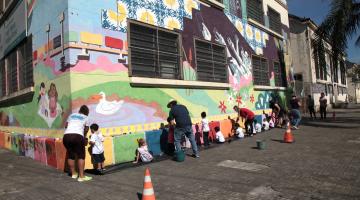 Fachada da escola pintada com vários motivos coloridos. Crianças e professores estão junto à parede finalizando o trabalho. #Pracegover