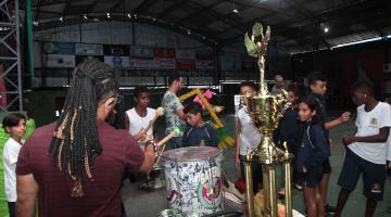 Estudantes estão na quadra da escola de samba em meio a troféus e instrumentos de percussão. #Paratodosverem