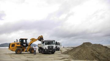 Escavadeira está depositando areia em caminhão. Os dois veículos estão na areia da praia. Ao lado direito da imagem há um monte de areia. #Paratodosverem.