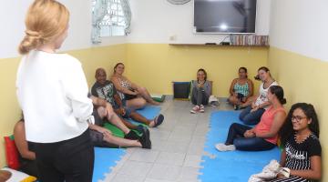 Mulher está em pé conveersando com adultos que estão sentados em colchonetes espalhados pelo chão. Na sala há uma tv e um ventilador. #Pracegover