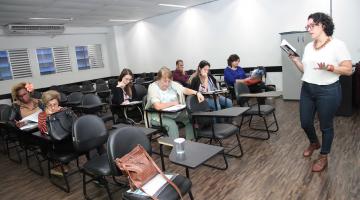 Professora com livro na mão fala para colegas em sala de aula #pracegover 