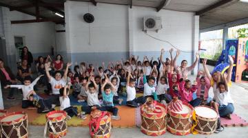 Crianças estão em pátio escolas, todas com as mãos para o alto. À frente do grupo há diversos tambores musicais. #Pracegover