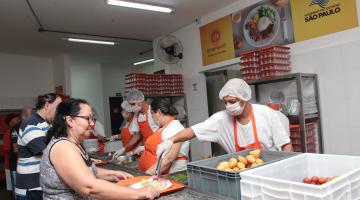 Pessoas estão à frente de balcão segurando bandejas. Do outro lado, pessoas com toucas e máscara servem as que seguram bandejas. #Pracegover