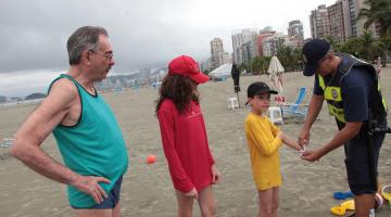 Guarda municipal coloca pulseirinha de identificação em criança na praia. Um adulto e outra criança estão ao lado. #Pracegover