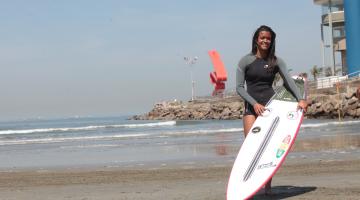 Surfista está na areia da praia segurança sua prancha. Ao fundo aparece o monumento de Tomie Ohtake no emissário submarino. #Pracegover