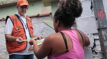 Agente da defesa civil usando colete e boné de identificação entrega folheto para moradores de área de risco. A imagem é fechada. Dá para perceber que os dois estão entre muros. #Paratodosverem