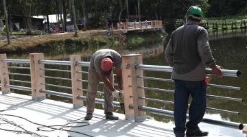 Dois trabalhadores estão instalando guarda-corpo na beira de uma lagoa. #Pracegover