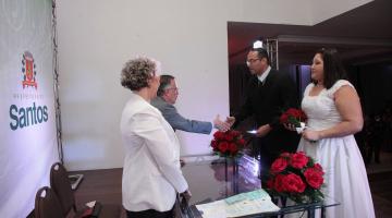 Casal de noivos, com as roupas tradicionais, está na frente de um casal atrás de uma mesa. Ao fundo um painel com o brasão de Santos. #Pracegover