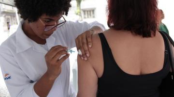 Mulher usando avental branco vacina o braço de outra mulher que está de costas. #Paratodosverem