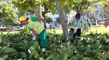 Pessoas uniformizadas, com pás e máscaras no rosto andam e manuseiam plantas em jardim. #Pracegover