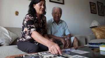 Amandia e Francisco estão sentados e apontam para fotografias que estão sobre uma mesa á frente deles. #Pracegover