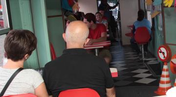 Passageiros estão dentro de carro de bonde onde se vê cones de trânsito e painel com orientações em geral sobre comportamento no trânsito. #Pracegover