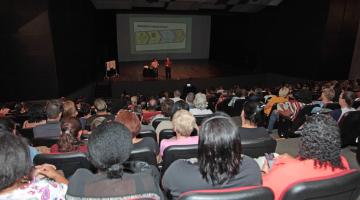 Teatro com cadeiras ocupadas majoritariamente por mulheres. Ao fundo, no palco, uma imagem está projetada em um telão. #Pracegover