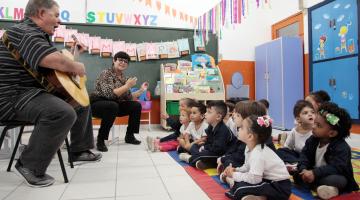 O músico está sentado tocando violão. Há uma professora ao lado dele. As crianças estão sentadas  no chão e acompanham. #Pracegover