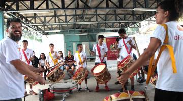 Jovens e adultos tocam instrumentos de percussão em pátio. #Pracegover