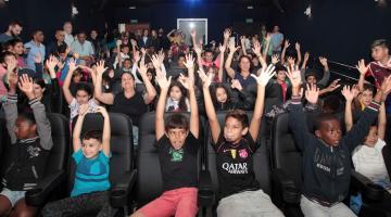 Crianças lotam sala de cinema. Todas estão sentadas nas poltronas e com as mãos para o alto, como se estivessem comemorando. #Pracegover