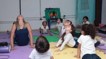 Crianças e professora fazem movimento no solo sobre tapetes de EVA. Elas estão com o corpo no solo e têm os dois braços esticados e paralelos. Todos flexionam a cabeça e olham para o alto. #Pracegover