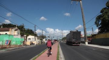 Via pavimentada com ciclovia ao centro. Ciclista pedala na ciclovia. Na pista de veículos à direita passa um caminhão. #Paratodosverem