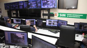 Sala de monitoramento das câmeras distribuídas pela Cidade. Há várias telas distribuídas pela sala. Dois homens estão assistindo. #Pracegover