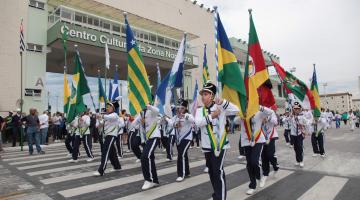 Estudantes uniformizados e segurando bandeiras percorrem avenida. #Pracegover