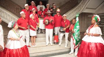 Integrantes de escola de samba estão na escadaria do hall do paço municipal. Eles vestem uniforme vermelho e branco. As mulheres estão com roupas típicas de baiana. #Pracegover