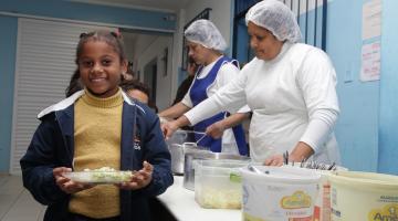 Criança caminha ao lado de mesa onde estão disponíveis os pratos com alimentos. Ela segura um prato com as duas mãos. #Pracegover