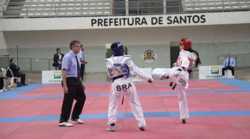 Duas atletas de taekwondo em luta sobre tatame. Ao lado esquerdo está o árbitro. #Pracegover