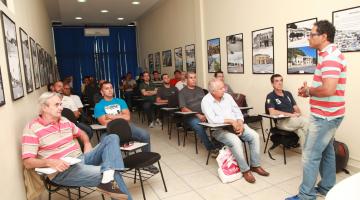 Sala de aula com várias pessoas sentadas ouvindo instrutor que está à frente de todos. #Pracegover