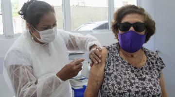 Enfermeira usando máscara aplica vacina em braço direito de uma mulher que também usa máscara #paratodosverem
