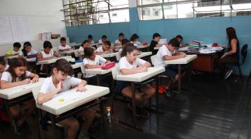 Alunos estão em sala de aula sentados e fazendo prova. Professora está no canto direito da imagem, com sua mesa próxima à parede da sala. #Pracegover