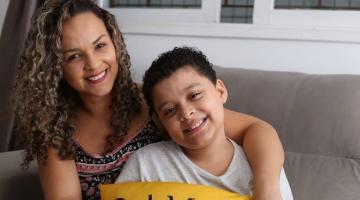 #pracegover no sofá de casa, Mariza e o filho Miguel posam sorridentes, abraçando almofada com a inscrição da palavra amor