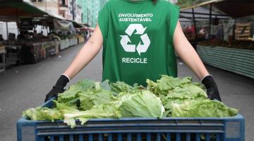 feira livrea ao fundo com destaque para mulher (sem aparecer o rosto) segurando engradado com verduras. Ela usa um colete onde se lê Envolvimento sustentável. Recicle. #paratodosverem 