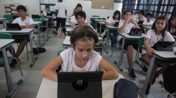 alunos estão sentados nas carteiras diante de tablets abertos. #paratodosverem